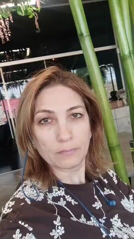 The video is a selfie taken by a woman with shoulder-length brown hair and light brown eyes. She is wearing a brown shirt with a white floral pattern and blue earbuds. Behind her, there are tall, green bamboo stalks. In the background, there are tables and chairs, suggesting an indoor setting like a restaurant or cafe. The woman is looking directly at the camera with a neutral expression. The video is short and appears to be a simple self-portrait.