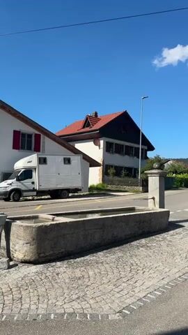The video shows a close-up of a water trough filled with water. Behind the trough is a street with a cobblestone sidewalk. A white truck with a trailer is parked on the street. There are houses and trees in the background. The sky is blue with a few white clouds.