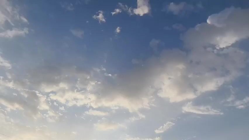 The video shows a view of a cloudy sky. The clouds are scattered across a bright blue sky, with some of the clouds appearing white and fluffy while others are darker and more gray. There are sunbeams visible, suggesting that the sun is either rising or setting. The overall impression is one of a dynamic and changing sky.