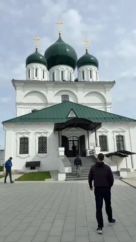 The video shows a plaza with several people walking in front of an orthodox church. The church has three green domes with golden crosses, white walls, and a green roof over the entrance. People are walking up the stairs to the entrance. The plaza is paved with grey tiles. Other buildings with white and blue walls and golden domes are visible in the background. The sky is overcast.