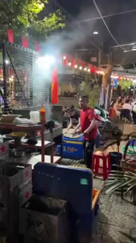 The video shows an outdoor restaurant in the evening. A man in a red shirt stands near a grilling station, gesturing to someone off-camera. Smoke rises from the grill. In the background, people sit at tables, eating and talking. Red flags and string lights are strung across the open-air space. There are coolers and other restaurant equipment scattered around. Vegetation is visible on the edges of the scene.