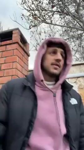 A man wearing a pink hooded sweatshirt and a black puffer jacket is outdoors near a brick chimney and a stone wall. He is speaking and looking around. The sky is overcast and there is a bare tree visible in the background.