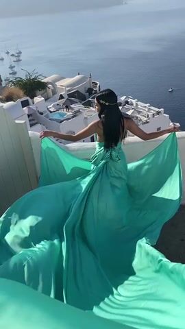 A woman with long dark hair, wearing a flowing teal dress and a gold headpiece, poses for a photo on a balcony overlooking a body of water. She is holding the train of her dress with both hands, her arms outstretched. In the background, there are white buildings with blue accents, a swimming pool, and sailboats on the water. The scene is bathed in bright sunlight.