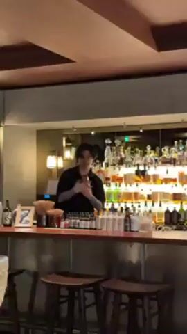 A bartender with dark hair and a black shirt is standing behind a bar, shaking a cocktail shaker. The bar is well-stocked with rows of bottles, illuminated from behind. There are several wooden stools in front of the bar. The ceiling has wooden beams.