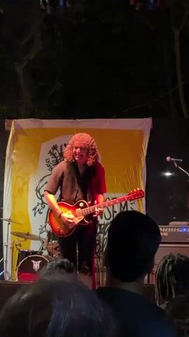 A man with curly brown hair is playing an electric guitar on a stage. He is wearing a black shirt and black pants. He is smiling and looking down at his guitar. In the background, there is a yellow banner with a drawing on it. To the left of the stage, there is a drum set. In the foreground, there are people in the audience. The scene appears to be a live music performance at night.