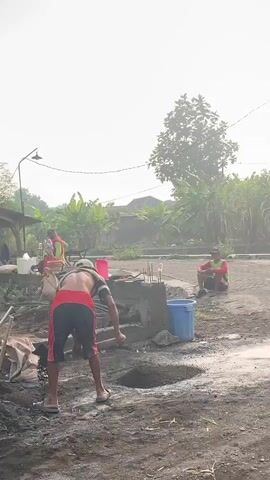 This is a video taken from a slightly elevated viewpoint, looking down at a construction site under a bright, overcast sky. In the foreground, a man wearing red and blue shorts, a white cap, and no shirt is bent over, seemingly mixing or moving a pile of dark construction material with a shovel. To his right, a blue bucket stands next to a pile of concrete. Further back, another man in red shorts and a green shirt sits on the ground. To the left, a woman in a red and white patterned top is working near some building materials and bags of sand. In the background, there are trees, including banana trees, and utility poles with wires. The ground appears to be dirt and wet in places. The overall atmosphere suggests an outdoor construction project in progress.