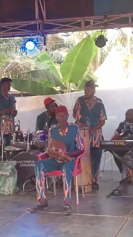 Here's a description of the video:

The video shows a live band performing on a stage. The band consists of a drummer, keyboard player, percussionists, and a lead singer who is speaking into a microphone. The musicians are all wearing matching outfits in dark teal with orange flame-like patterns. The lead singer is wearing a red suit and cap. The stage is covered by a metal structure with a red roof, and there are large banana trees behind the stage. The video seems to be taken at an outdoor event. The band's equipment is visible, including a drum set labeled "Victory Drums" and a keyboard with "YAMAHA EFAC" on it. The stage floor appears to be tiled or covered with a material that has a tile-like pattern.  There are speakers and other stage equipment around. People are seated at tables in the background.