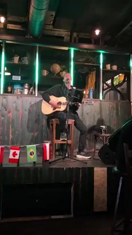 A man sits on a stool on a small stage, playing an acoustic guitar and singing into a microphone. He wears a black sweater and dark pants. A laptop is positioned on a stand in front of him, and various flags are displayed on a rope in front of the stage, including flags of Canada and Brazil. The background features a wooden wall with green lighting. The scene appears to be an indoor venue like a bar or small club.
