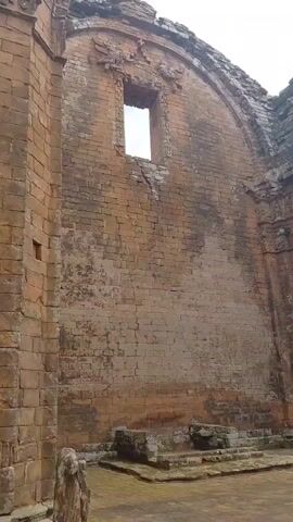 The video shows the ruins of an old brick structure, possibly a church or mission. The walls are made of red brick and show signs of age and decay, with patches of missing mortar and weathered surfaces. There are remnants of arches and doorways, as well as a single small, rectangular window in one of the walls. The floor appears to be paved with stone or brick. In the foreground, there is a low, square stone structure with steps leading down into it. The environment suggests an outdoor historical site, with a cloudy sky overhead and some trees visible in the distance. The overall impression is one of ancient grandeur in a state of ruin.
