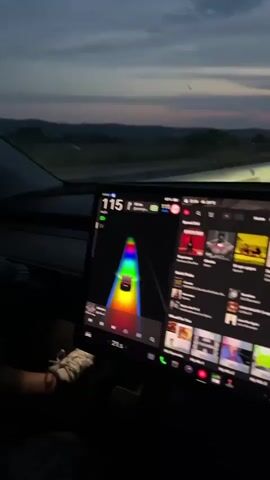 The video is shot from the interior of a car, facing forward through the windshield during dusk or nighttime. A large digital display on the car's dashboard occupies the lower half of the frame. The left side of the display shows a car's driving interface with a speed of "115" and a colorful, rainbow-like visualization of the road ahead, indicating lanes or surroundings. The right side of the display shows a media player interface with multiple album covers or video thumbnails on a dark background. Beyond the windshield, a multi-lane highway is visible, illuminated by the car's headlights and distant streetlights. The sky is dark with faint remnants of a sunset on the horizon. The car appears to be in motion.