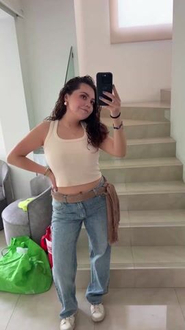 A young woman is taking a mirror selfie in a hallway. She is wearing a light beige tank top, light blue jeans with a brown fringe belt, and white sneakers. She is holding a black smartphone in her right hand and has her left hand on her hip. Her hair is curly and dark, and she is wearing jewelry. Behind her is a staircase with white treads. To the left are a gray upholstered chair, a bright green shopping bag, and a red bag. The hallway is well-lit with white walls and tiled floors. The mirror has a metallic frame. The camera zooms out slightly during the shot.