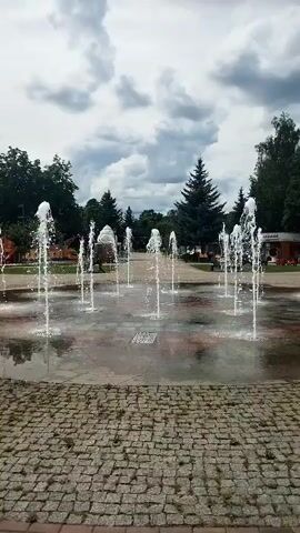The video shows a dynamic water fountain display in an outdoor park setting. Several jets of water erupt from the ground in a synchronized manner, creating a visually engaging spectacle. The fountain is situated on a paved area with a textured stone border in the foreground. In the background, lush green trees and a cloudy sky contribute to the serene atmosphere. A few people can be seen in the distance, adding a sense of human presence to the scene. The overall impression is one of a lively and refreshing public space.