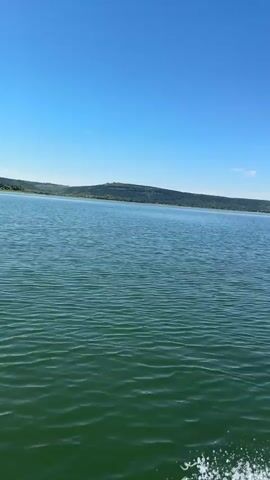 A boat is moving on a large body of water. The water is green and choppy, with white spray visible at the edge of the frame, indicating movement. The sky is a clear, bright blue with a few wispy white clouds in the distance. The horizon is lined with a green, hilly landscape. The camera is pointed forward and slightly to the right, capturing the view from the boat.