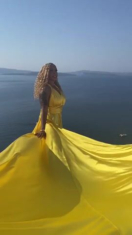 A woman with blonde braided hair stands on a ledge overlooking the ocean. She is wearing a yellow halter dress that flows out around her. The woman is looking to her left and smiling. The ocean is blue and calm, with islands in the distance. The sky is clear and blue. The video is taken from a low angle, looking up at the woman and her dress.