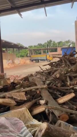 A pile of cut logs and branches fills the foreground of the video. Behind the logs, a dirt yard is visible, with a yellow truck parked in the distance. A covered structure with a metal roof extends across the top of the frame, and in the background, there are trees and partially constructed buildings. The sky is cloudy. The video is short and there are no people visible.
