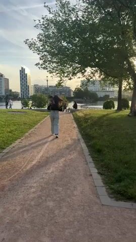 A woman walks away from the camera on a gravel path in a park. She is wearing a black top and light blue jeans and is carrying a child in her arms. The path curves to the left and is bordered by a grassy area on the right. In the background, there is a body of water, and beyond that, several modern buildings. Sunlight filters through the leaves of a large tree on the right side of the frame. Other people can be seen in the distance sitting on the grass and walking. The overall scene appears to be a park setting on a sunny day.