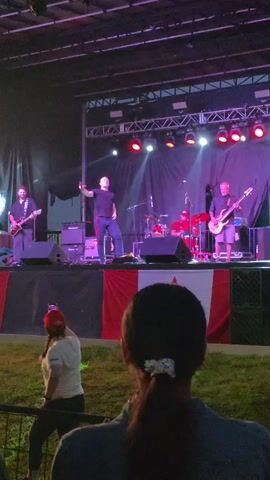 The video shows a live music performance on an outdoor stage. A band of four men is performing, with one vocalist in the center, two guitarists on either side, and a drummer behind. The stage is lit with purple and red lights. A Canadian flag is displayed prominently on the front of the stage. In the foreground, the back of a woman's head and shoulders are visible as she watches the performance. She is wearing a red helmet and holding a phone up, presumably to record the event. The audience is seated on grass in front of the stage.