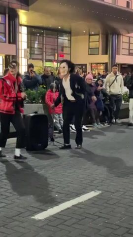 Two street performers dressed in costumes are dancing in front of a crowd of onlookers. The first performer is dressed in a red jacket and black pants, reminiscent of Michael Jackson, and is holding a microphone. The second performer is wearing a black suit and a creepy mask. They are dancing on a brick-paved street, with a black speaker behind them. The crowd is a mix of ages, and some are recording the performance with their phones. The background includes modern buildings with large windows and shops. It appears to be a public square or pedestrian area.