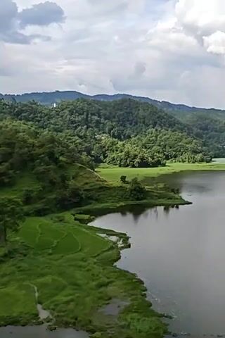 The video opens with a wide shot of a serene lake surrounded by lush green hills. The water is calm, reflecting the cloudy sky and the verdant landscape. As the camera pans, it reveals terraced fields along the shoreline, adding to the picturesque scenery.

The perspective then shifts to a long, suspension bridge made of metal. The bridge stretches across a vast expanse, connecting two densely forested hills. The camera moves forward along the bridge, offering a view of the dense greenery that blankets the slopes.

The final scene provides a panoramic view of the lake from the bridge, with the vibrant green hills continuing to dominate the horizon under a dynamic sky filled with white clouds. The video captures the natural beauty of the location, emphasizing the tranquility of the lake and the grandeur of the surrounding hills and forests.