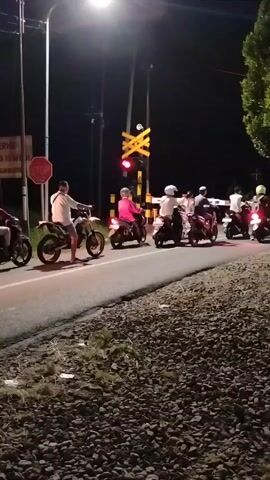 A group of motorcyclists are stopped on a road at night, possibly waiting for a train. The road is paved and bordered by gravel. A railroad crossing signal with a red light is visible in the background. There are streetlights and trees in the scene.