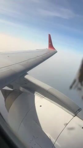 The video shows an airplane wing and engine, viewed from inside the plane. The sky is visible in the background, with a light blue color above and a hazy white layer below. The wing is gray, and the winglet is red. The engine is white with some grime and detailing. There is a slight blur from the window. The plane is flying.