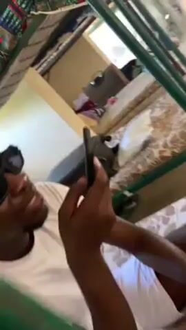A dark-skinned man wearing sunglasses and a white shirt is lying down and talking on a cell phone. He is looking directly at the phone as he speaks. The background includes a bunk bed above him and a suitcase nearby.