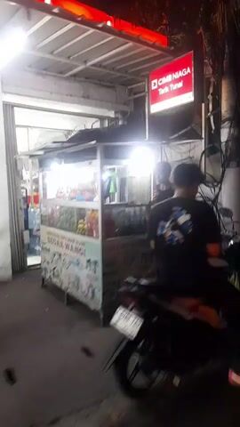 The video shows a street scene at night, likely a food stall or market. A person in a dark shirt is seen working at a stall with bright lights. Another person in a light-colored shirt and green pants is also visible near a food cart. There are various objects and structures, including a red bucket, food displays, and overhead lighting. Indonesian flags are visible in the background. The scene appears to be bustling with activity, suggestive of a lively commercial area.