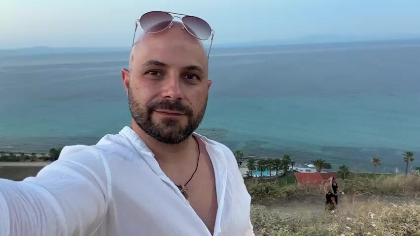 This is a selfie video of a bald man with a beard, wearing a white open-collared shirt and sunglasses on his head. He is standing on a grassy hillside overlooking a turquoise body of water. In the background, there are some buildings with swimming pools, palm trees, and other vegetation. Two women, one in a black dress and the other in dark clothing, are visible in the middle ground, walking down a dirt path. The sky is overcast and appears to be either early morning or late evening, suggesting dawn or dusk. The overall mood of the video appears relaxed and vacation-like. There is no visible text in the video. The location is identified as Kassandra Municipal Unit, Greece.