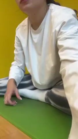 A young woman is stretching on a green yoga mat. She is wearing a long-sleeved white shirt, shiny gray leggings with a swirl pattern, and white socks. She is sitting with her legs spread in a wide straddle and bending to her right side, her left arm extended overhead. The background is a bright yellow wall. The camera is positioned low, capturing the woman from a low angle.