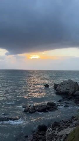 The video shows a rocky coastline with the sea in the foreground and a stormy sky in the background. The sun is setting or rising, and its light is visible through a gap in the clouds, casting a reflection on the water. The waves are gently crashing against the rocks. There are no people or animals visible in the video. The overall mood is serene and slightly dramatic due to the stormy sky.