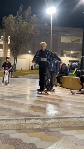 At night, the video shows three males skateboarding on a paved, outdoor plaza, in front of a large building with lighted windows. The plaza is designed with large hexagonal tiles in a patterned color scheme.

The first male, blond-haired, dressed in a black track suit, performs a skateboard trick, a 360 flip. He makes the landing successfully.

The second male, standing nearby to the right, wears a blue jacket and is riding his skateboard slowly, facing right.

The third male stands further in the background to the left, with his skateboard.

There are several people sitting on benches in the background. A tree stands to the left of the scene. Tall lightposts illuminate the area.