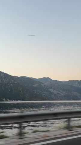 This is a first-person perspective video taken from a vehicle moving along a road next to a body of water. The road has a guardrail separating it from the water. In the background, there are mountains. The sky is clear and light blue, suggesting it is either early morning or late afternoon. The water is calm and reflects the sky and mountains. There are no people visible in the video.