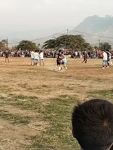 The video shows a soccer match being played on a dirt field. A crowd of spectators is gathered around the field. There are two teams of players, one wearing white and blue striped jerseys and the other wearing black and white jerseys. The players are actively engaged in the game, with one player from the black and white team attempting to tackle a player from the white and blue team. The ball is visible in the air at one point. The background shows a hilly landscape with some trees and buildings. The sky is overcast. The video is shot from a low angle, with the heads of two spectators visible in the foreground.
