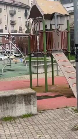 The camera pans around a playground. It's an overcast day, and there are apartment buildings in the background. Two children, dressed in dark jackets, are playing on the playground equipment. One child is on a playground structure with a slide, and the other is running near a different slide. The playground has various structures, including slides, swings, and climbing equipment. The ground is a mix of green grass and red flooring. A concrete block is visible in the foreground. There are several parked cars visible in the background.