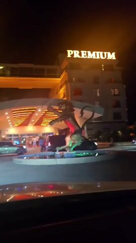 The video shows the entrance to a hotel and casino at night. A large statue of a winged lion is in the foreground, with a fountain around its base. The hotel building is illuminated with golden lights, and the word "PREMIUM" is displayed in large letters at the top. The entrance to the hotel features a covered walkway with red lighting. Several cars are parked in front of the entrance, and people can be seen walking into the building. The overall impression is one of a luxurious and lively establishment.