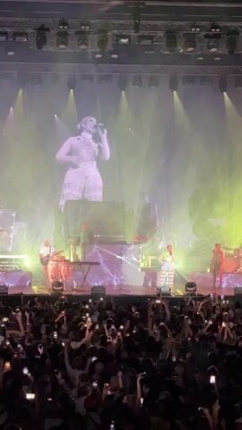 The video shows a live concert with a large audience in the foreground. On stage, musicians are performing. A large screen behind the performers displays a projection of two women, one playing a cello and the other a violin. The lighting is dramatic, with spotlights and colored lights illuminating the stage and creating atmospheric haze. The audience is holding up their phones to record or take pictures of the performance.