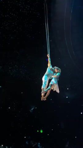 A man and woman, both wearing white outfits, hang from a long, thin, white rope suspended from the ceiling of a large, dark auditorium. They are performing an acrobatic aerial act. The man holds the rope with one hand and the woman with the other as he balances on one leg in a split position. The woman hangs off the man, extending herself horizontally. The auditorium's ceiling is covered in twinkling lights resembling stars. Some audience members are visible at the bottom of the frame.