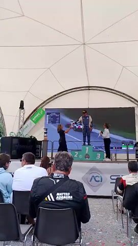 A man in a racing suit stands on the center podium (labeled 1) holding a trophy. Two women are on either side of the podium, one on the left (stage right) handing him the trophy, and one on the right (stage left) standing next to him. A large screen behind them displays a race scene. The podium has numbers 1, 2, and 3 displayed. Below the podium are logos for ACI and S. In the foreground, people sit in chairs facing the stage. The scene appears to be outdoors under a white tent-like structure.