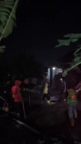 The video captures a nighttime scene where several individuals, many wearing high-visibility vests and yellow boots, are engaged in what appears to be a clean-up or rescue operation. The environment is dark, illuminated by artificial light sources, suggesting it's either nighttime or an indoor scene with limited lighting. The ground is wet and muddy, indicating recent flooding or water damage. People are holding tools, possibly shovels or brooms, and seem to be working together. The presence of a vehicle with a red light also suggests an ongoing operation. The overall atmosphere suggests a situation requiring collective effort to address a problem.