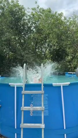 The video shows a person in an above-ground swimming pool. The pool is blue and has a silver ladder with white handrails. Green trees and foliage are visible in the background. The person is in the water, splashing, and appears to be smiling. The water is blue and appears to be disturbed, indicating movement or activity. The sky is cloudy.