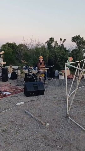 The video shows a four-member band performing outdoors on a gravelly surface. The band consists of a drummer at the back, a guitarist/vocalist at the front, a singer on the left, and a bassist behind the guitarist. The stage area is marked by a rug. The equipment includes two speakers, amplifiers, and various musical instruments. A small group of people are sitting behind the band, acting as an audience. The background features trees and a dusk sky. The scene is cluttered with cables and miscellaneous objects.