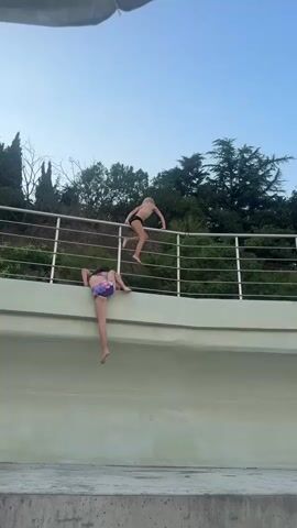Two children are on a railing above a concrete ledge, possibly the edge of a pool. The child closer to the camera, wearing a purple and pink patterned swimsuit, is hanging by their arms from the railing, with their legs dangling down. The second child, further up the railing and to the right, is wearing black swim trunks and is crouched on the railing, appearing to be preparing to climb over or jump. The background consists of trees and a clear sky. The overall scene suggests playful or daring activity.