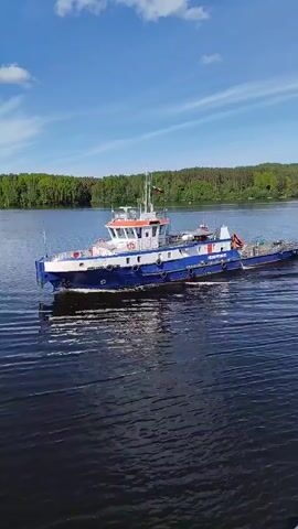 The video captures a medium-sized blue and white vessel, likely a workboat or tugboat, actively moving across a body of water. The boat features a distinct white superstructure with an orange-colored roof on its upper deck, and a blue hull with a white band running along its side. Black fenders are visible along the hull's perimeter.

As the vessel moves, it generates a clear white wake behind it on the dark blue water. The sky overhead is bright blue with scattered white clouds, indicating a clear and sunny day. In the background, a continuous line of dense green trees forms the distant bank of the waterway, suggesting a natural, possibly forested, landscape.

On the side of the boat's hull, the word "РОСМОРПОРТ" (ROSMORPORT) is clearly visible in white capital letters. A small flag, possibly the Russian national flag, can be seen flying from a mast on the boat's cabin. The scene depicts a calm and operational maritime setting, likely on a river or large lake near Nikol'skaya Gora, Russia.