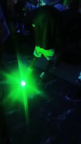 The video shows a dimly lit indoor setting, possibly a club or party, with several people moving in the background. The scene is dominated by a green and then blue light source that casts strong, starburst-shaped rays on the dark floor. The floor appears to have a marble or textured pattern. One person, seen from behind, is wearing dark clothing, light-colored shorts that glow under the light, and a hat. This individual is moving or dancing. Other people are visible in the background, but their details are obscured by the low lighting.