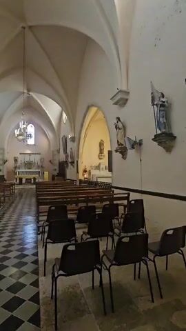 The video shows the interior of a church. In the foreground, there are several rows of dark chairs. Behind the chairs, there are wooden pews. The floor in the foreground has a black and white checkered pattern. The walls of the church are white and are decorated with religious statues and artwork. The ceiling is arched with ribbed vaults. Further into the church, an altar can be seen at the far end. The overall impression is a peaceful and solemn atmosphere.