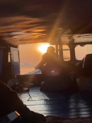 The video shows the interior of a boat with wooden flooring and a dark ceiling. The sun is setting or rising, casting a warm orange glow. Two people are visible. One person, silhouetted, is sitting facing away from the camera, towards the light. Another person's arm and shoulder are in the foreground on the left, also facing away from the camera. There are some objects on the floor, including shoes and bags. The overall atmosphere is calm and serene, with the light creating a beautiful backdrop.