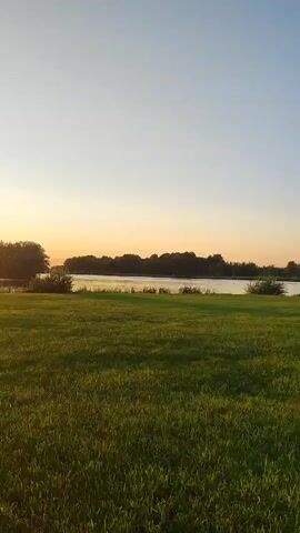 A still shot of a sunset over a grassy field and a body of water. The sun is setting on the left side of the frame, casting a warm glow across the sky and the water. Trees line the far bank of the water. The grass in the foreground is green and lush. The sky is clear with a gradient of colors from orange and yellow near the horizon to blue at the top of the frame.