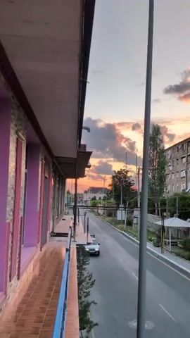 The video captures a street scene in Şəki, Azerbaijan, during a vibrant sunset. The camera is positioned on a balcony overlooking a road, with a building in the foreground on the left. The building's facade is a mix of stone and purple-colored panels, with a tiled balcony floor.

The street is asphalted and appears to be in a residential or semi-commercial area. A silver car is visible driving away from the camera. Power lines and utility poles crisscross the view, indicating an urban or semi-urban setting. In the background, several multi-story buildings and houses are present, suggesting a town or city. Lush green trees add to the scenery.

The most striking element is the sky, which is illuminated by the setting sun. Dramatic clouds are painted with hues of orange, pink, and gray, creating a picturesque and serene atmosphere. The time of day is clearly sunset, given the warm, dramatic lighting and the colors in the sky. The weather appears clear, with the exception of the colorful clouds.

No people are directly visible in the frame. The overall mood conveyed is peaceful and visually captivating, thanks to the beautiful sunset. There is no visible text in the media file.