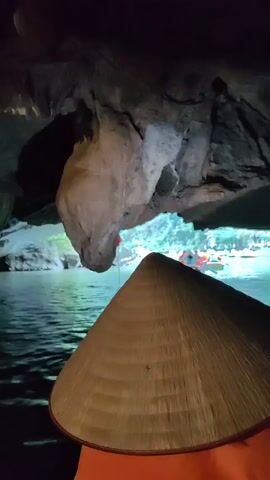 The video shows a person in a boat entering a dark cave. The person is wearing a traditional Asian conical hat. The boat is traveling on water that reflects the light. The cave has a rocky ceiling and walls. There is a red flag visible in the distance outside the cave. Other boats are visible in the distance.