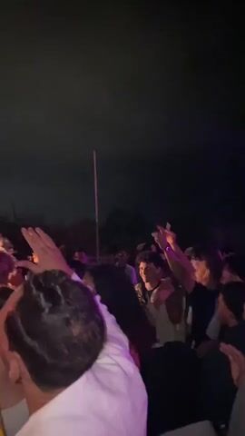 A nighttime outdoor gathering is underway. A DJ stands behind equipment, wearing headphones and a sleeveless shirt. A man in a light-colored jacket stands to the DJ's left, holding a drink. Another man in a red shirt stands behind the DJ. A crowd of people is visible, many with their arms raised. The sky is dark and appears to be clear. There are trees and some structures in the background. The lighting is dim and focused on the DJ area.
